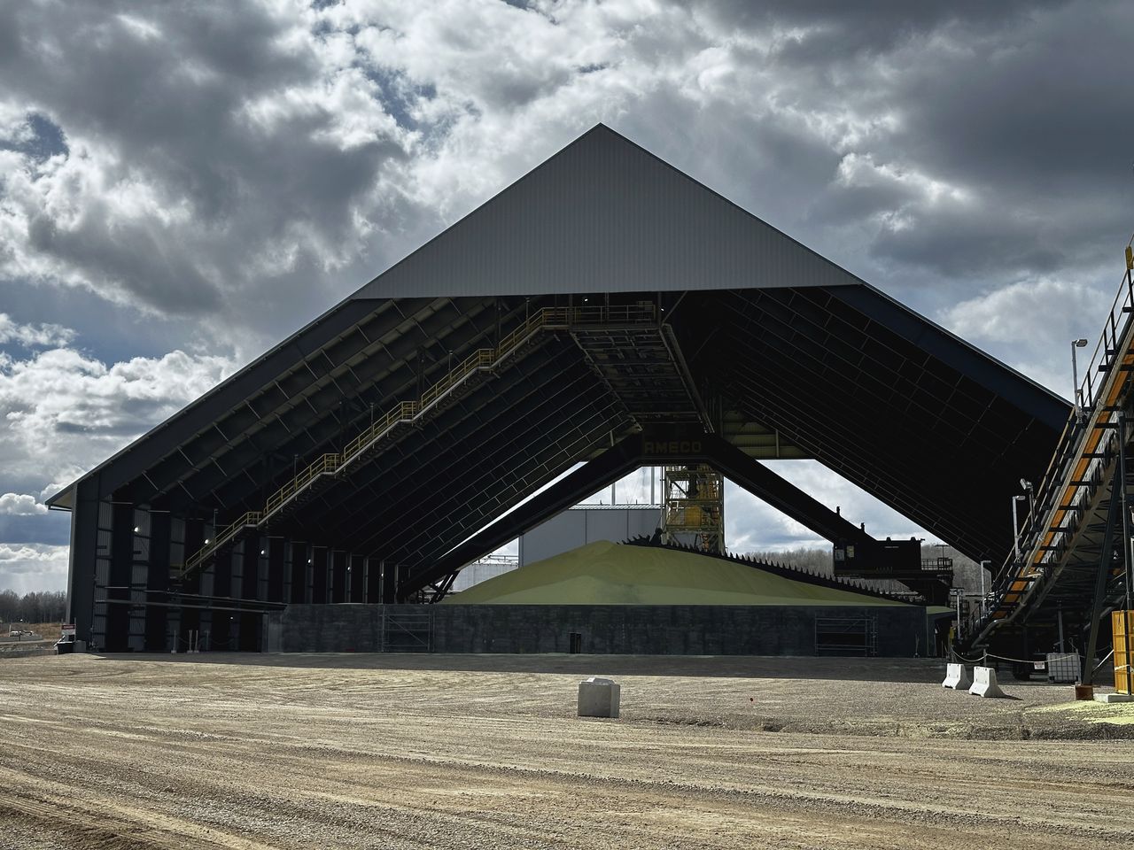 A-frame bulk storage building under a dramatic prairie sky beside a rail-served terminal